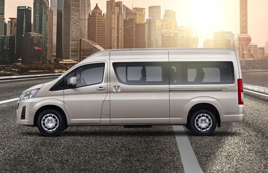 Minibus Toyota Hiace berwarna krem, tampak samping, dengan latar gedung kota
